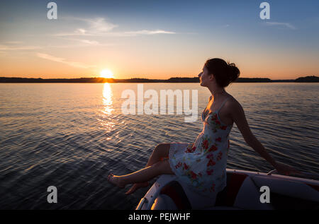 Jeune femme assise sur le bord d'un bateau sur un lac au coucher du soleil. Banque D'Images