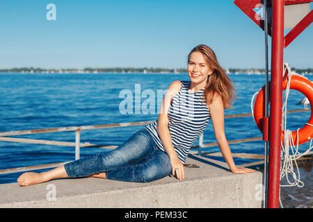 Beautiful smiling white Caucasian girl femme avec de longs cheveux blonds portant des jeans et t-shirt à rayures de la mer assis dehors sur le lac bright sunny s Banque D'Images