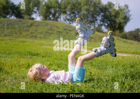 Les loisirs, l'enfance, des jeux de plein air et sport concept - Jolie petite blonde la petite fille se trouve sur l'herbe en patins à roulettes Banque D'Images