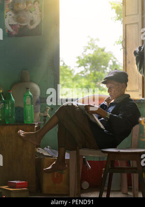 Un vieil homme birman est assis sur une chaise et lit un journal dans le soleil du matin. Banque D'Images