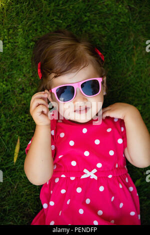 Cute little girl se trouve sur l'herbe sous le soleil de l'été les lunettes. Banque D'Images