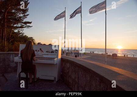 Jurmala, Lettonie. Août 13, 2018. Une jeune femme joue du piano dans le coucher de soleil à la plage de la mer Baltique de Lettonie Jurmala. Avec une vue panoramique sur la mer, les passants peuvent jouer un piano peint par des artistes sur la promenade près de la salle de concert Dzintari. Crédit : Alexander Welscher/dpa/Alamy Live News Banque D'Images