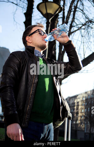 Beau jeune homme avec des lunettes debout sous les lumières dans le parc, l'eau potable à partir d'une bouteille et la pensée. Banque D'Images