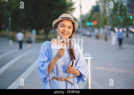 Portrait of beautiful happy smiling laughing Caucasian blanche fille brune femme avec la peau tannée en robe bleue et hat eating ice-cream cone, coucher du soleil Banque D'Images