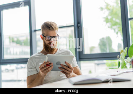 Le multitâche. Beau jeune homme portant des lunettes et de travail avec surface tactile tout en étant assis sur le canapé dans office Banque D'Images