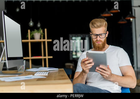 Le multitâche. Beau jeune homme portant des lunettes et de travail avec surface tactile tout en étant assis sur le canapé dans office Banque D'Images