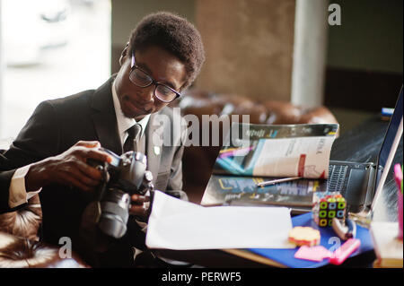 African American man photographe paparazzi de l'usure sur un costume noir et de lunettes assis au bureau de travail avec l'appareil photo et ordinateur portable derrière. Banque D'Images