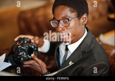 African American man photographe paparazzi de l'usure sur un costume noir et de lunettes assis au bureau de travail avec l'appareil photo et ordinateur portable derrière. Banque D'Images