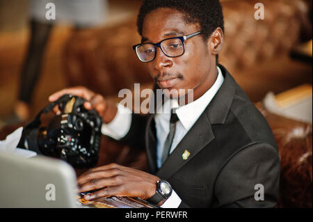 African American man photographe paparazzi de l'usure sur un costume noir et de lunettes assis au bureau de travail avec l'appareil photo et ordinateur portable derrière. Banque D'Images