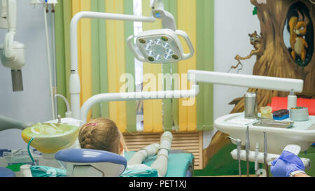 Enfant avec une mère à la réception d'un dentiste. La jeune fille se trouve dans le fauteuil, derrière sa mère. Le médecin travaille avec un assistant. Procédure pour le forage d'un dent. La fixation d'un joint temporaire. Banque D'Images