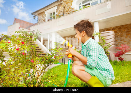 Vue côté-portrait de jeune garçon de sept ans à l'aide de gicleurs d'arrosage rosiers main dans le jardin Banque D'Images