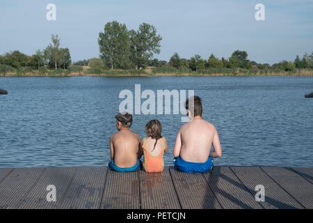 Des enfants assis sur la jetée. Trois enfants d'âge différent - adolescents, l'âge préscolaire et primaire garçon fille assise sur une jetée en bois. L'été et l'enfance concept. Les enfants sur un banc au bord du lac. Frères et soeurs assis sur la jetée en bois Banque D'Images