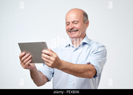 Smiling mature male using digital tablet contre fond gris. Trouver l'information nécessaire dans le web. Étude pour l'utilisation d'internet au cours de la vieillesse Banque D'Images