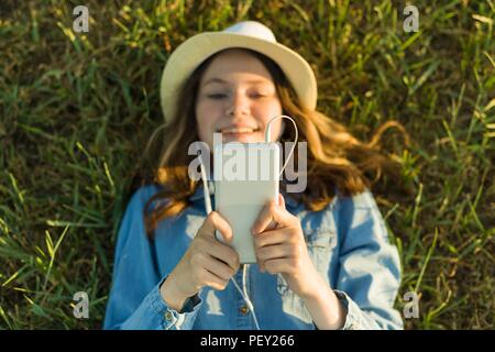 Teenage girl in hat avec des écouteurs se trouve sur l'herbe verte et ressemble au téléphone. L'accent sur smartphone, vue d'en haut Banque D'Images