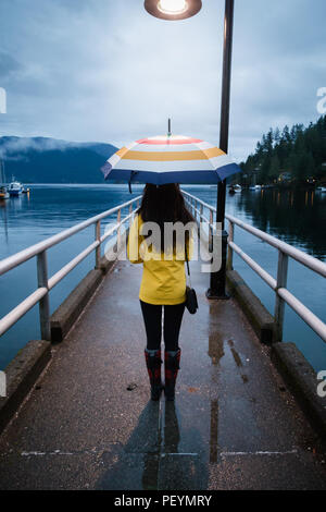 Jeune femme dans un imperméable jaune permanent, sous un parapluie un jour de pluie par l'océan. Banque D'Images