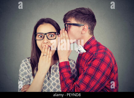 Jeune homme en secret à dire lunettes femme à choqué en se tenant sur le fond gris Banque D'Images