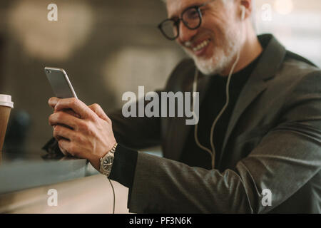 Mature businessman with earphones assis au café comptoir avec smart phone dans la main. Se concentrer sur les hommes hand holding cell phone. Banque D'Images