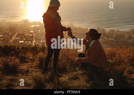 Jeune homme à genoux et proposant femme avec fleur sauvage sur la montagne. Belle vue sur la montagne en proposant deux avec la lumière du soleil et du paysage marin Banque D'Images