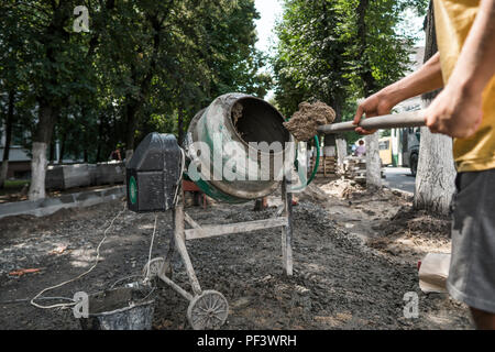 Travailleur de la construction Ajouter les ingrédients pour le mélange à la bétonnière sur chantier à l'aide d'une pelle pendant les travaux de construction de trottoirs Banque D'Images