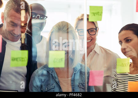 Groupe diversifié de smiling businesspeople standing in a modern office ensemble un remue-méninges avec les notes sur un mur de verre Banque D'Images