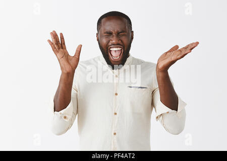 Emotive excité African American guy avec barbe en chemise blanche de crier à haute voix de bonheur et d'émerveillement, gesticulant avec palmiers, fermer les yeux, d'être très joyeux et heureux sur mur gris Banque D'Images
