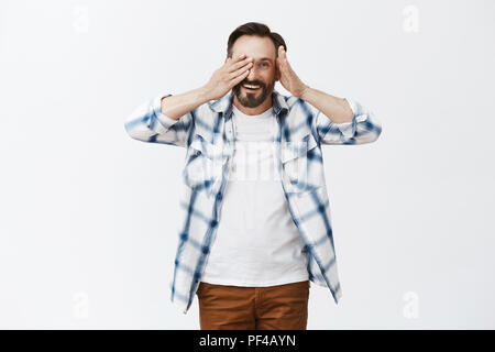 Peek-a-boo cutie. Portrait de l'environnement et genre beau mâle mature avec barbe, décollant de Palm et de l'oeil regarder joyeusement de l'appareil photo, jouer avec les enfants s'amusant sur fond gris Banque D'Images