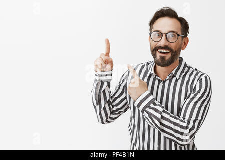 Incroyable, regardez ici. Portrait de charmé et impressionné emotive caucasian man with beard dans les verres et chemise rayée, pointant dans le coin en haut à gauche et souriant joyeusement, posant sur mur gris Banque D'Images