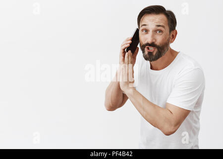 J'ai besoin de réponse, appel important désolé. Portrait de l'homme charmant occupé entrepreneur avec barbe, holding smartphone près de l'oreille et couvrant l'orateur en fléchissant vers la caméra et murmurer quelque chose Banque D'Images