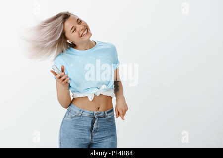 Portrait of cute woman excité positive à bretelles, sauter et danser, s'amuser et profiter d'un son des écouteurs sans fil, écouter la musique et holding smartphone, souriant avec les yeux fermés Banque D'Images
