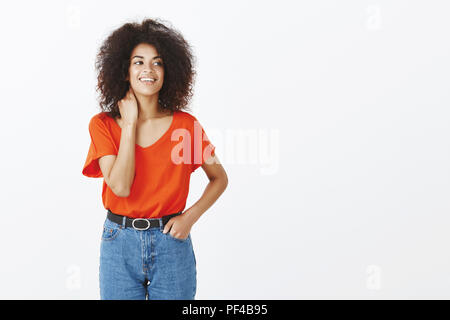 Tourné à l'intérieur de belle femme à la peau sombre avec la coiffure afro de toucher le cou et à droite avec charmant sourire, se sentant peu nerveux avant d'importante réunion sur mur gris Banque D'Images