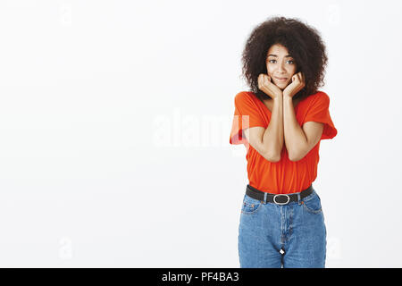 Portrait de l'insécurité timide aux cheveux bouclés attrayant étudiante en costume élégant, tenant les mains sur les joues et souriant de l'adjudication d'expression, d'être timide et amical sur mur gris Banque D'Images