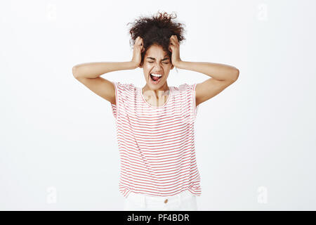 Ne peut pas contenir ses émotions à l'intérieur. Portrait of a souligné la peau sombre emotive femme en t-shirt à rayures, tenant les mains sur la tête, fermer les yeux et criant à haute voix, d'être dans la dépression et en colère sur mur gris Banque D'Images