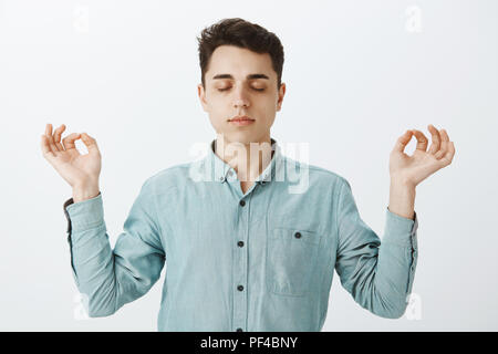 Taille plan sur l'accent calme gars en chemise, soulevant les mains dans le zen geste, fermer les yeux et de méditer, pratiquer le yoga ou le bouddhisme, se sentir détendu et heureux sur mur gris Banque D'Images