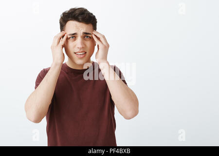 Portrait d'malheureux gars en t-shirt rouge, touchign les tempes avec les doigts et froncer, souffrant de migraine ou de maux de tête, mécontent et permanent sur fond gris sombre Banque D'Images