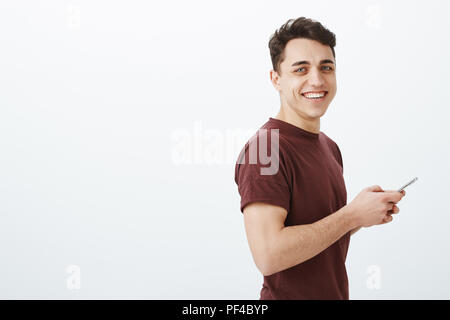 Photo de profil amical positif dans l'petit ami occasionnels t-shirt, holding smartphone et contemplant avec large sourire à l'appareil photo, profiter de passer du temps en ligne et en parlant avec des amis sur mur gris Banque D'Images