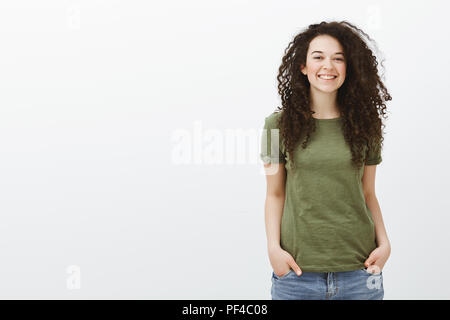 Portrait of cute timide étudiante aux cheveux bouclés dans dark-t-shirt vert, tenant les mains dans les poches et sourire tout en se sentant à l'aise et d'allégresse en nouvelle société sur mur gris Banque D'Images