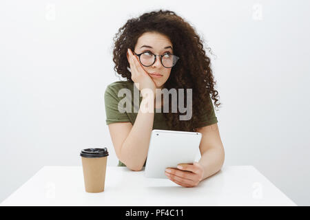 Ennuyer assez mignonne petite amie à lunettes, assis à table, la tête penchée sur le côté droit et a fixer indifféremment, boire du café et holding digital tablet, sensation de l'ennui sur fond gris Banque D'Images