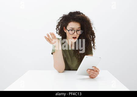 Frustrés choqué Belle aux cheveux bouclés creative designer lunettes très tendance en noir, assis à table, raising hand cluelessly et confus à tablette numérique, voir nouvelles décevantes Banque D'Images