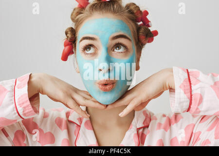 Close-up portrait of european girly girl avec expression mignon, vêtu de pyjamas et bigoudis, debout avec le masque facial sur fond gris, tenant la main sur le menton et à des lèvres plissées. Banque D'Images