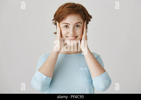 Je ne suis pas à l'écoute, honnêtement. Portrait of attractive young positives'femme aux cheveux rouges et de rousseur couvrant les oreilles avec des paumes et souriant joyeusement, debout sur fond gris Banque D'Images