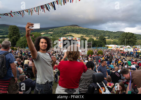 Glanusk Park, Brecon, pays de Galles, 18 août 2018. Deuxième jour du festival de musique Green Man dans les montagnes Brecon Beacons au pays de Galles. Un jeune homme cool prend un selfie devant la scène principale et la montagne à l'approche d'une tempête de pluie. Plans généraux de la foule et du site pendant le festival – scène principale de montagne. Crédit : Rob Watkins/Alamy Live News Banque D'Images