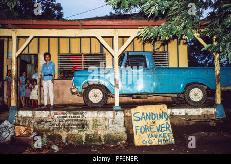 Managua, Nicaragua, juin 1986 ; une femme a juste stationné par pick-up par sa maison. L'enseigne prend en charge les sandinistes contre une possible invasion soutenue par les Etats-Unis de l'autre côté de la frontière du Honduras. Banque D'Images