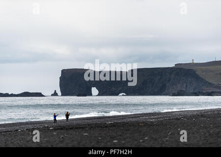 Islande Vik, deux jeunes femmes touristes sur black sand beach, ocean horizon, rock formation arch à Dyrholaey, Kirkjufjara photographier le phare sur so Banque D'Images