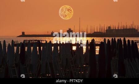 Belle pleine lune rouge au-dessus du port vu de la plage avec parasols Banque D'Images