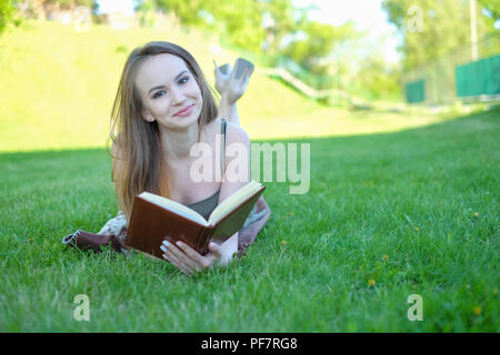 Jeune femme se trouve sur l'herbe verte et lit d'adresses dans le parc de la ville. Banque D'Images