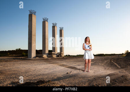 Déçu femme debout au milieu de la construction routière et de la tenue d'un tournesol cassé pendant le coucher du soleil. La destruction de l'environnement concept. Banque D'Images