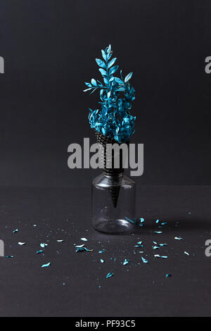 Des brindilles peintes en bleu clair avec peu de feuilles bleu dans une gaufre en corne noire dans un verre sur un papier noir. Banque D'Images