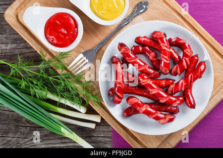 Saucisses grillées sur une assiette blanche avec de la sauce tomate et la moutarde dans une sauce bateaux sur une planche à découper sur un tapis de table, petits oignons et l'aneth sur un vieux wo Banque D'Images