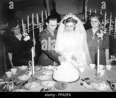 Photographie noir et blanc, montrant un groom dans un uniforme militaire et verres, aider une femme vêtue d'une robe blanche, voile, Tiara, et les verres, de couper un gâteau de mariage, avec des gâteaux, de l'argenterie et des bougies sur la table devant eux, et à lunettes, les femmes d'âge moyen de chaque côté, photographié dans l'Ohio, 1945. () Banque D'Images