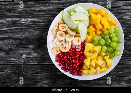 De délicieux fruits colorés - banane, pomme, mangue, ananas coupé en tranches et morceaux, les raisins et les graines de grenat sur plaque blanche sur l'ancien onglet rustique foncé Banque D'Images